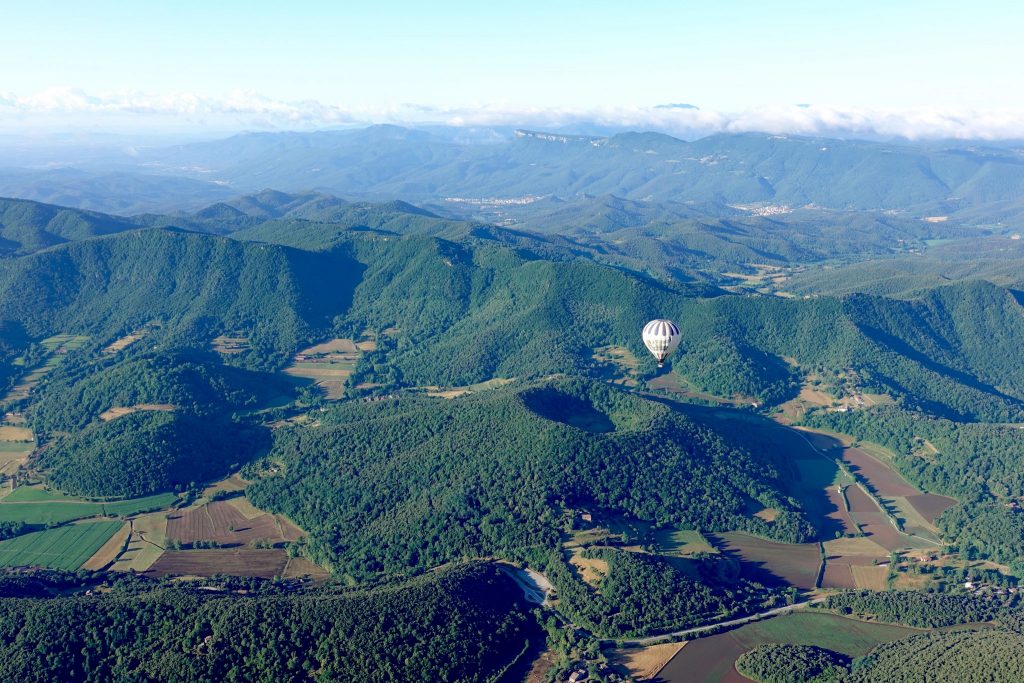 Blick auf die grüne Landschaft, einen bewachsenen Kraterkegel der Garroxta von oben, ein Heißluftballon steigt gerade auf