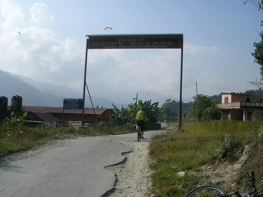 Ich fahre mit dem Rad durch einen Torbogen mit der Beschriftung "Happy Village", am Himmel sind Paragleiter zu sehen, die aspahltierte Straße ist zum Teil gebrochen und fehlt